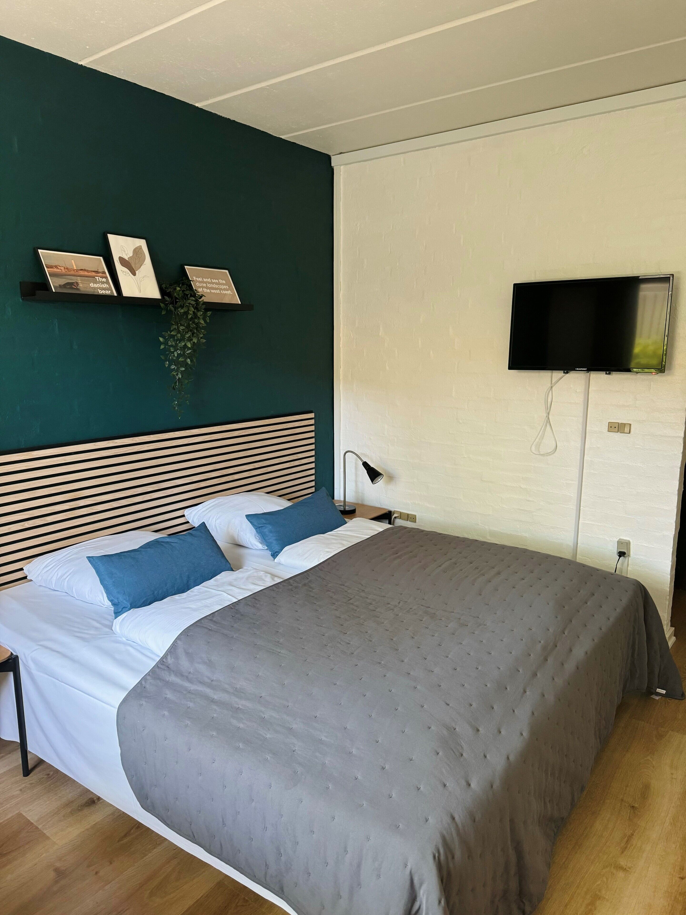 A neatly made bed with a grey duvet, a wooden headboard, and blue pillows. A flat-screen TV is mounted on the wall to the right.