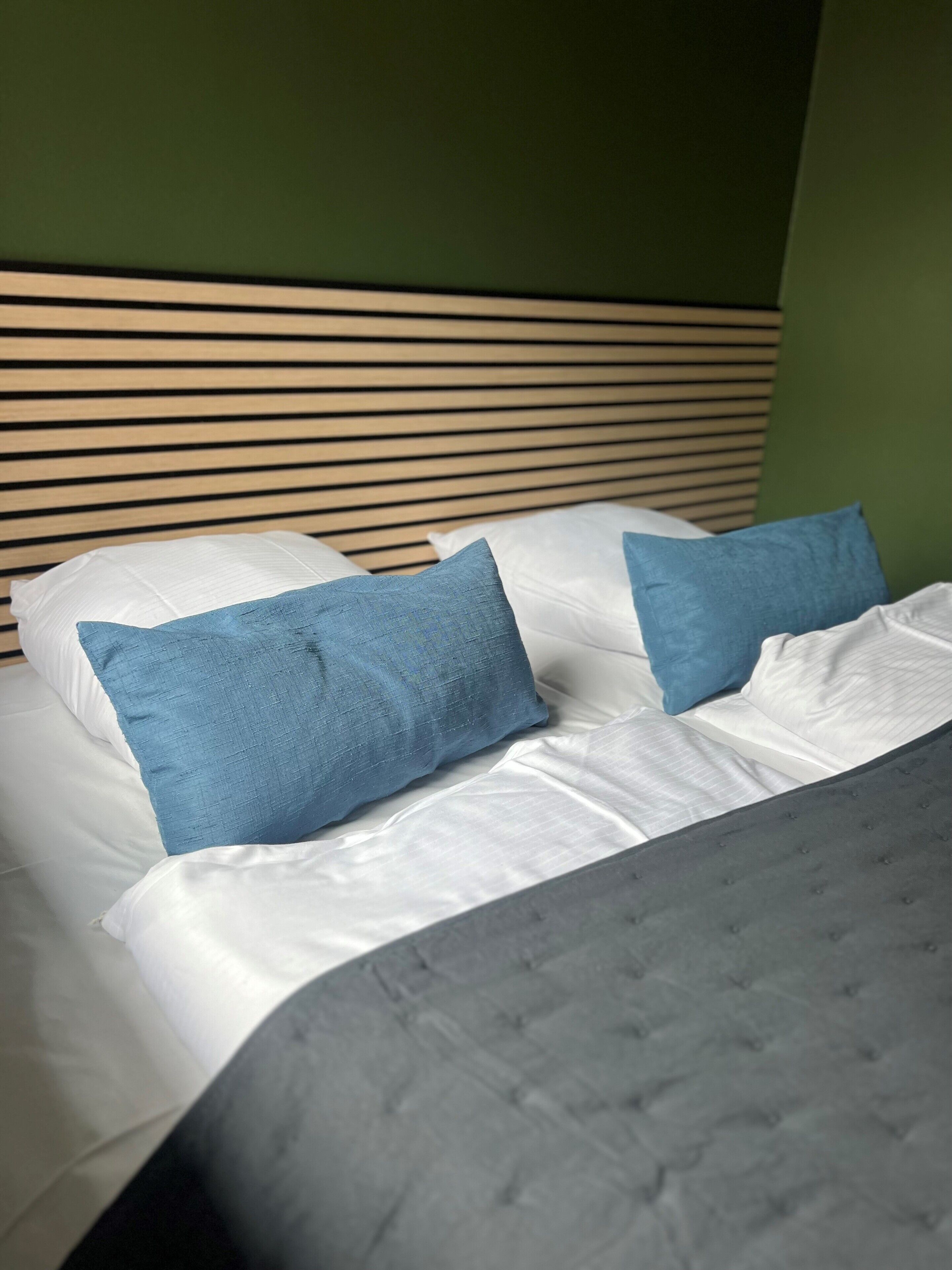 A bed with white linens and two blue pillows, a grey upholstered headboard, and a green wall.