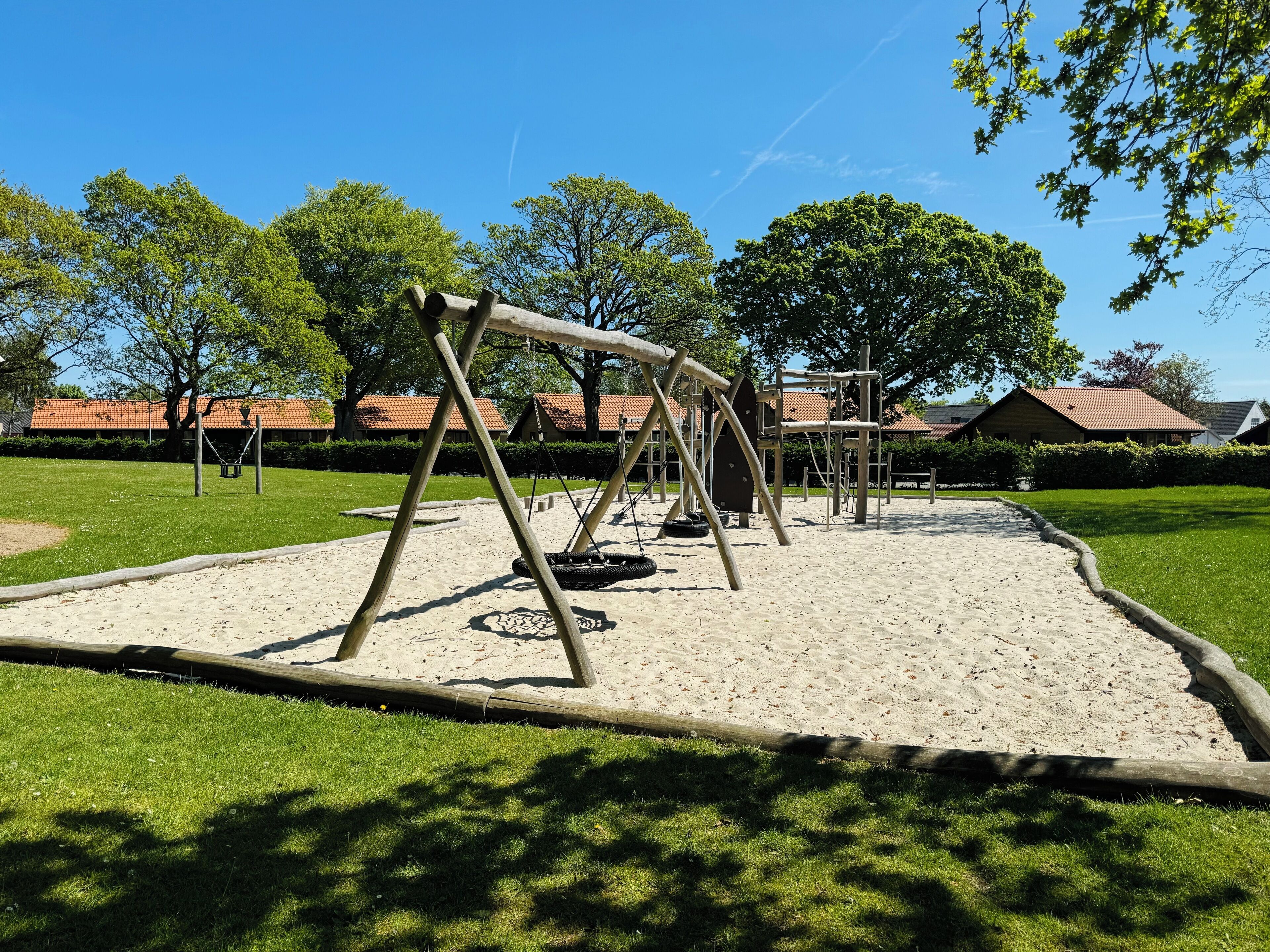 Un parque infantil con columpios y un neumático colgante, rodeado de césped y árboles.
