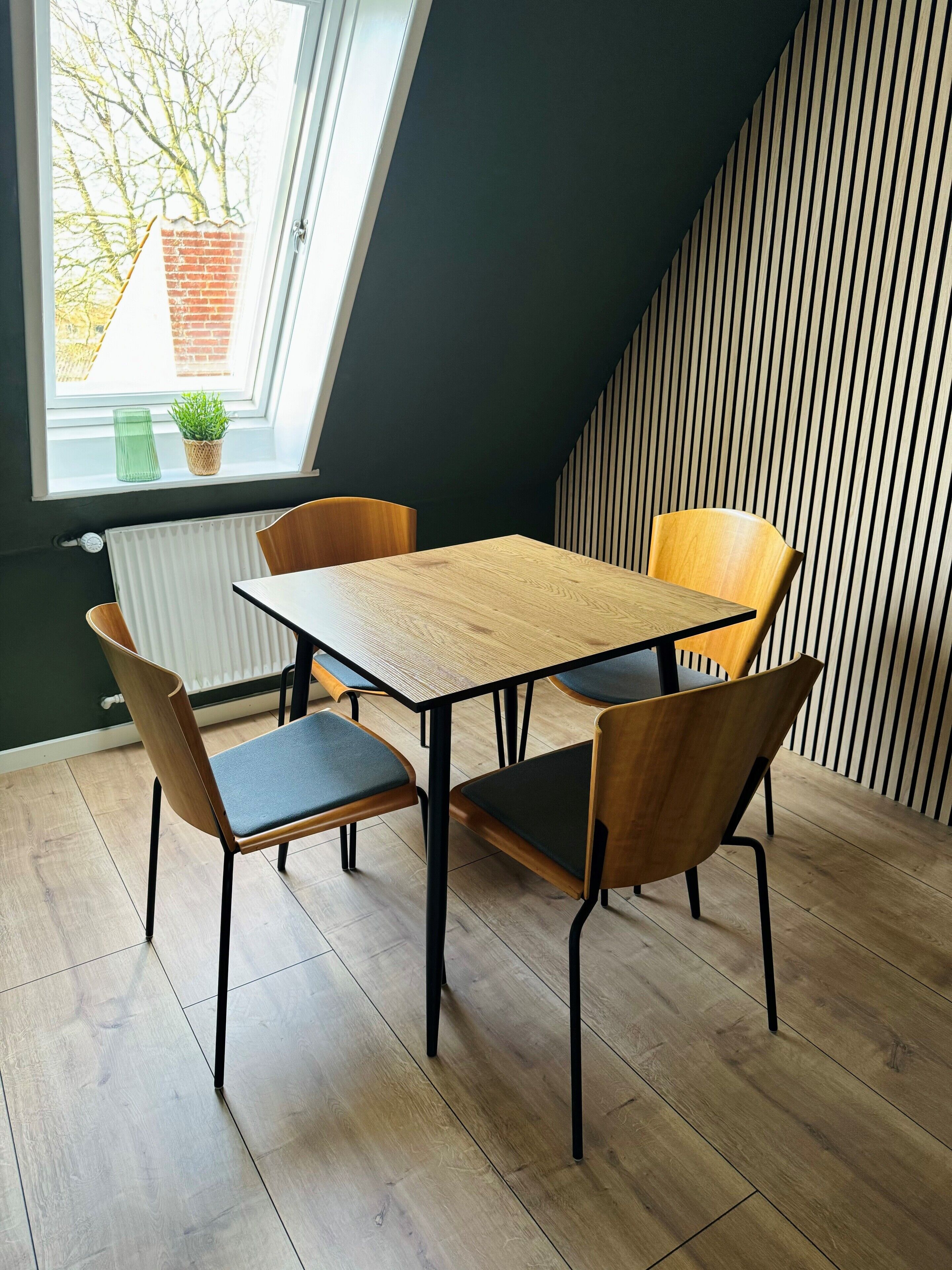 Een zithoek met een tafel en vier stoelen, een raam met uitzicht op een bakstenen gebouw en een radiator aan de muur.