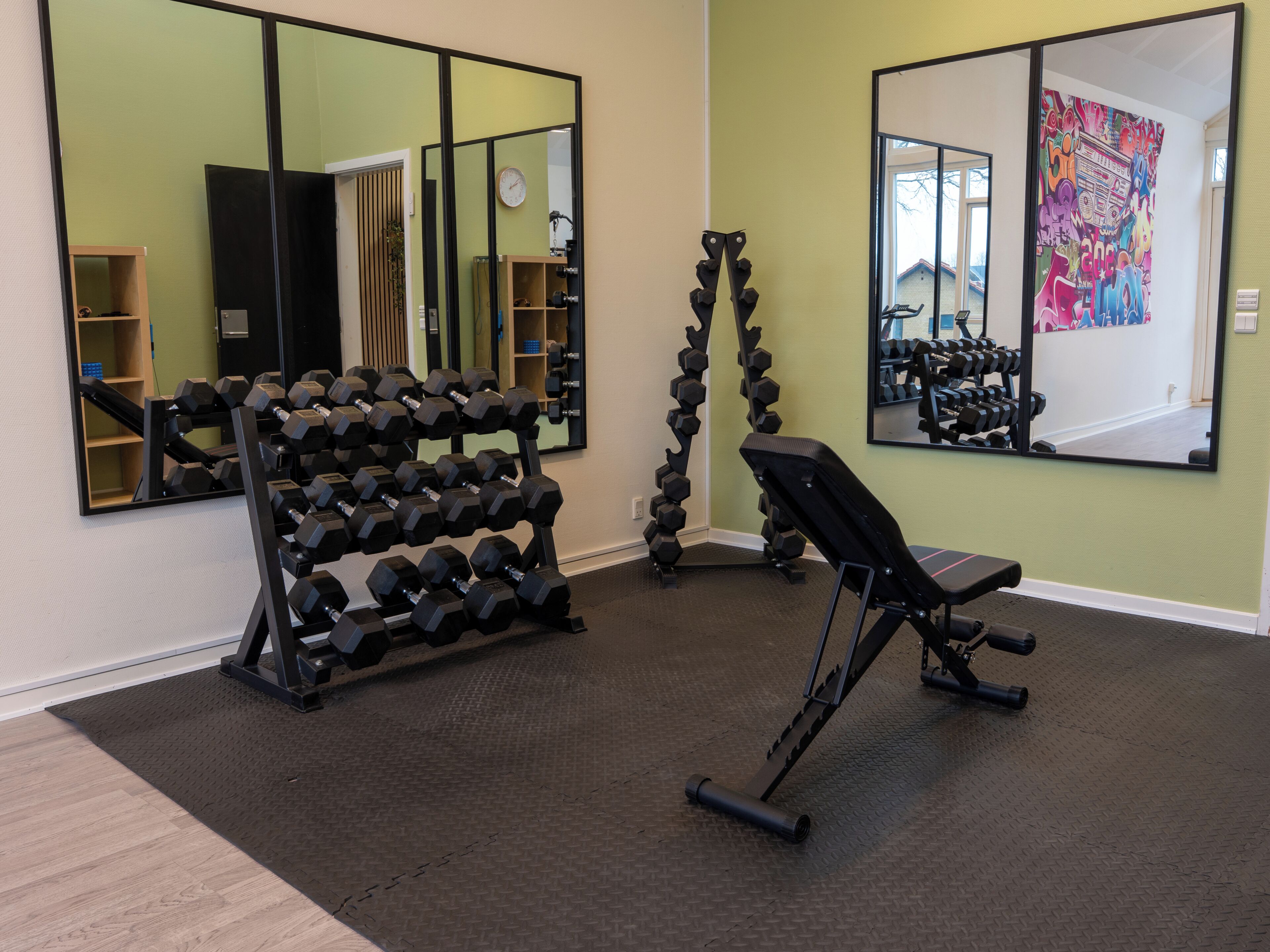 Un gimnasio con equipo de ejercicio, espejos y una pared decorada con una obra de arte colorida.