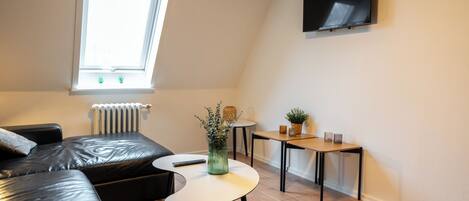 Un salon moderne avec un canapé en cuir noir, une petite table ronde, une plante en pot et un téléviseur à écran plat fixé au mur.