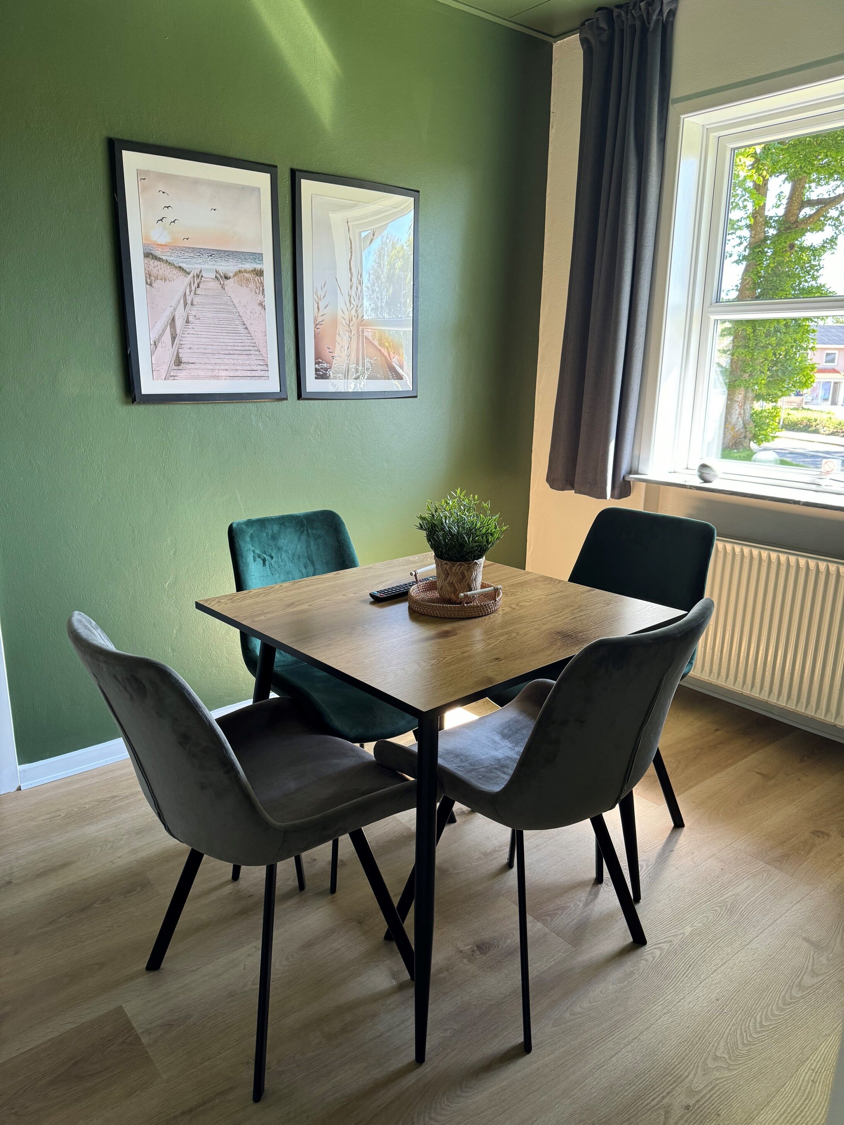 Un comedor con una mesa de madera, cuatro sillas, dos cuadros en la pared, una planta en maceta y una ventana con cortinas.