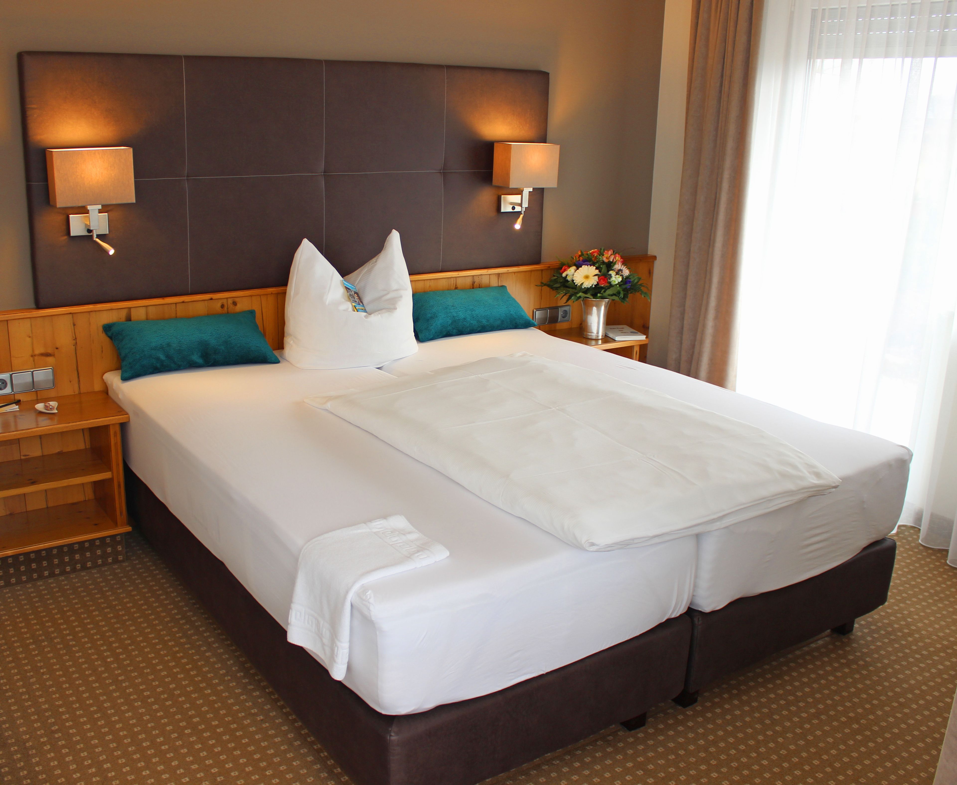 A hotel room with a large bed, two bedside tables, a nightstand, a headboard with wall-mounted lamps, and a window with curtains.