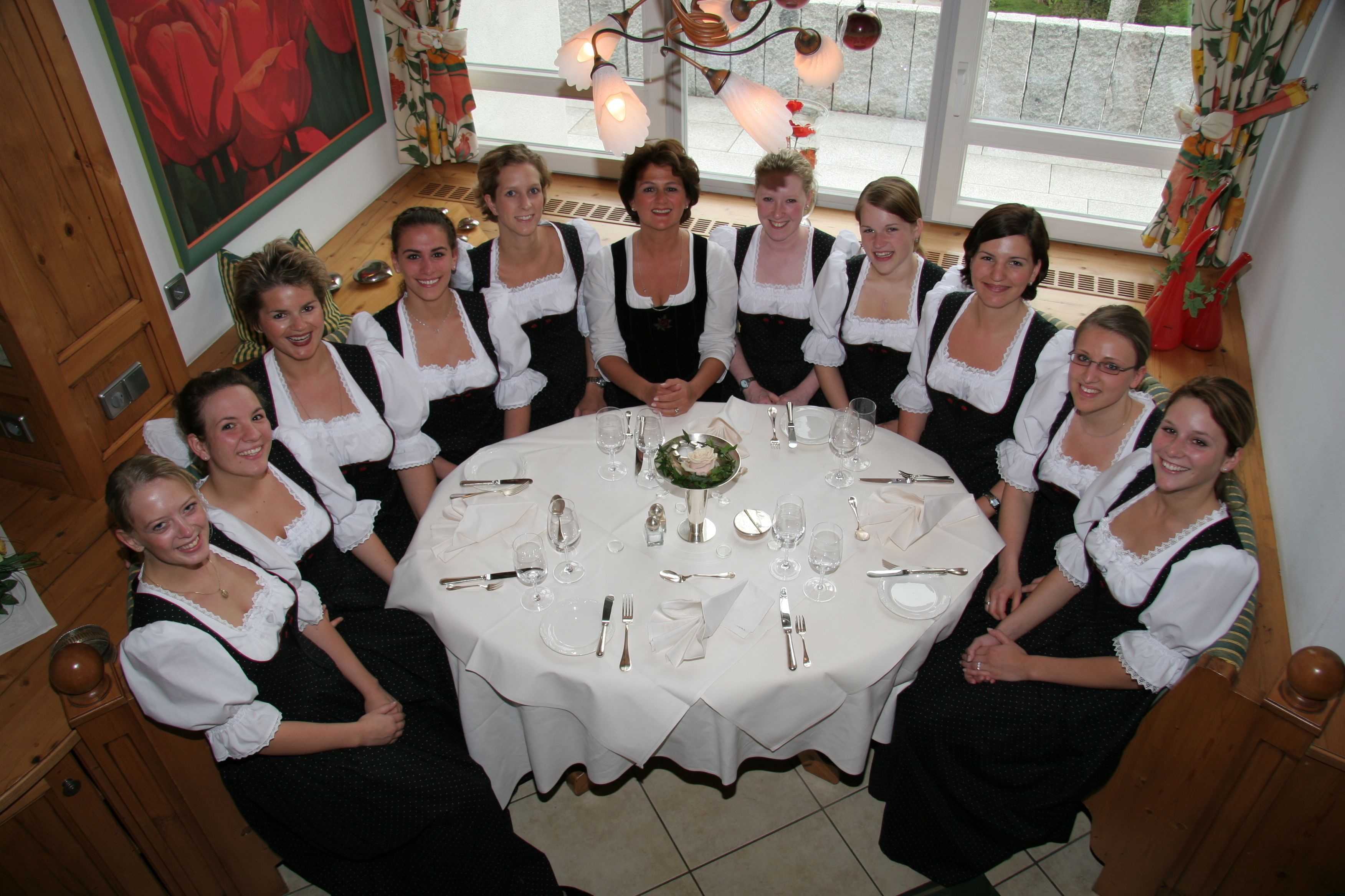A group of people seated around a round table with a white tablecloth, in a room with wooden floors and a large window with curtains.