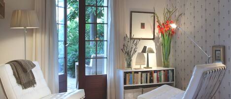 A bedroom with a large window, a white sofa, a bedside table with books, a lamp, and a vase with flowers.
