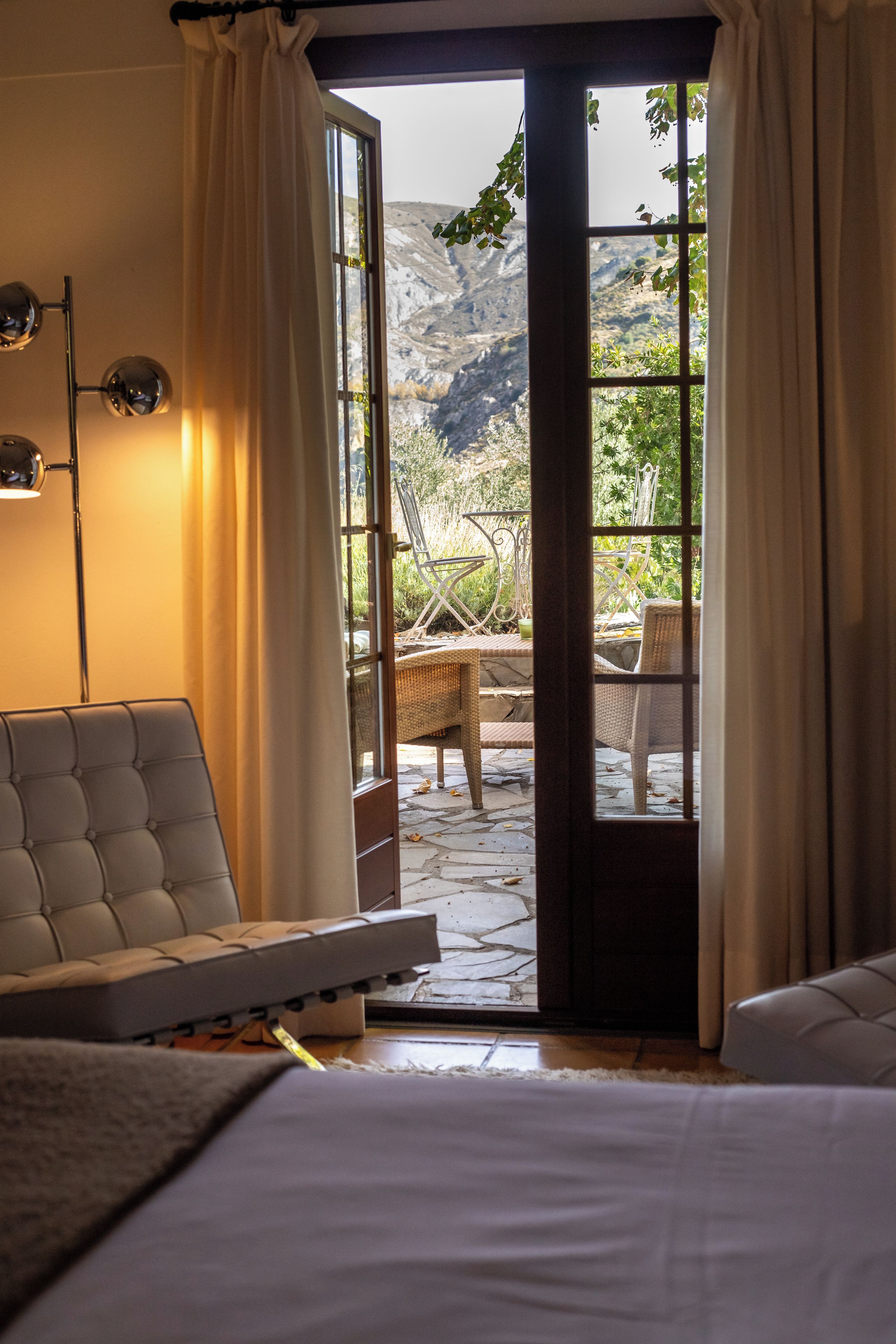 A room with a bed, a chair, and a table with a view of a mountainous landscape through a glass door.