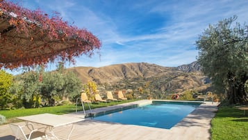 Piscina com espreguiçadeiras, pergolado com folhagem avermelhada e montanhas ao fundo.