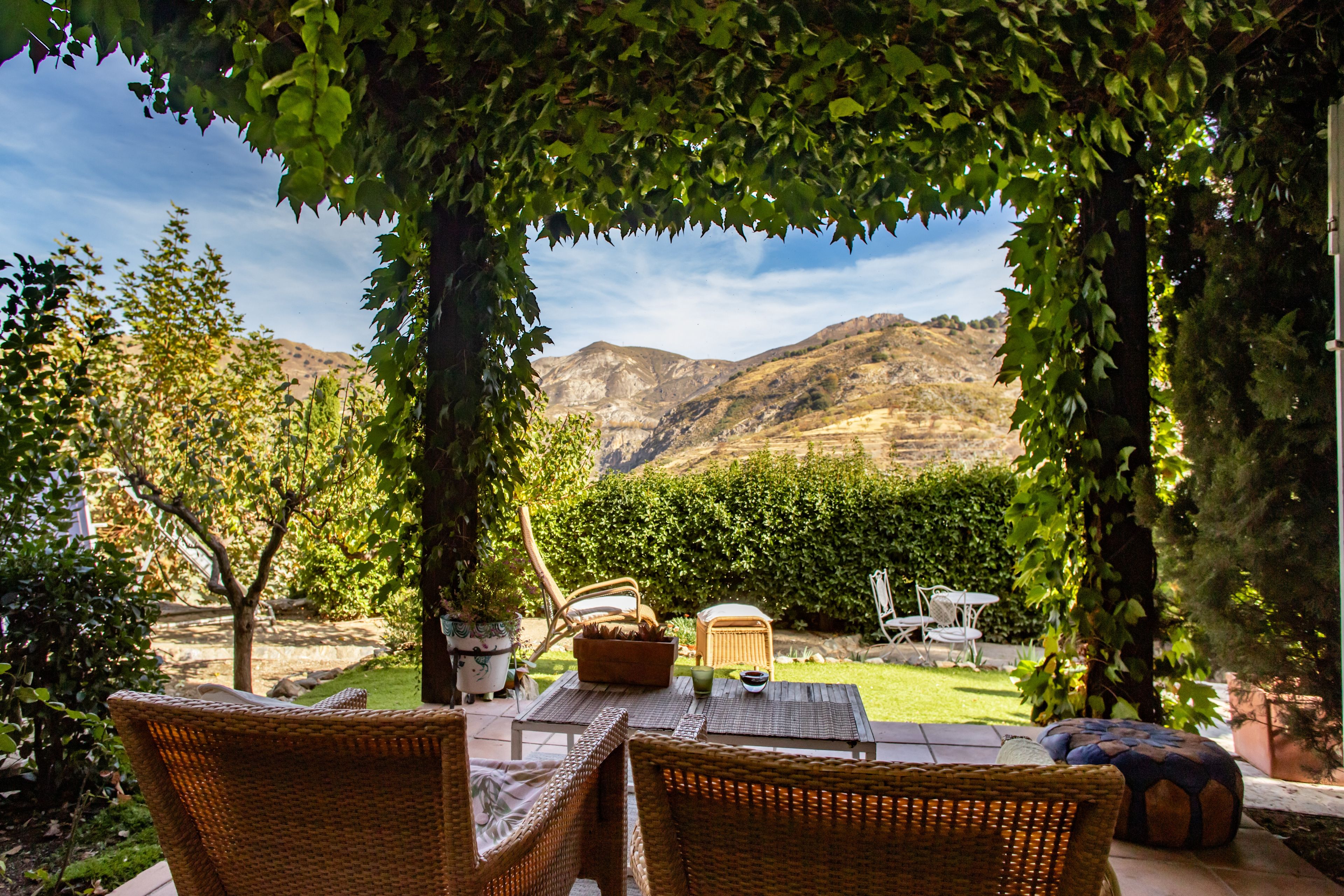 An outdoor seating area with wicker furniture, a table, and chairs, overlooking a landscaped garden and mountains.