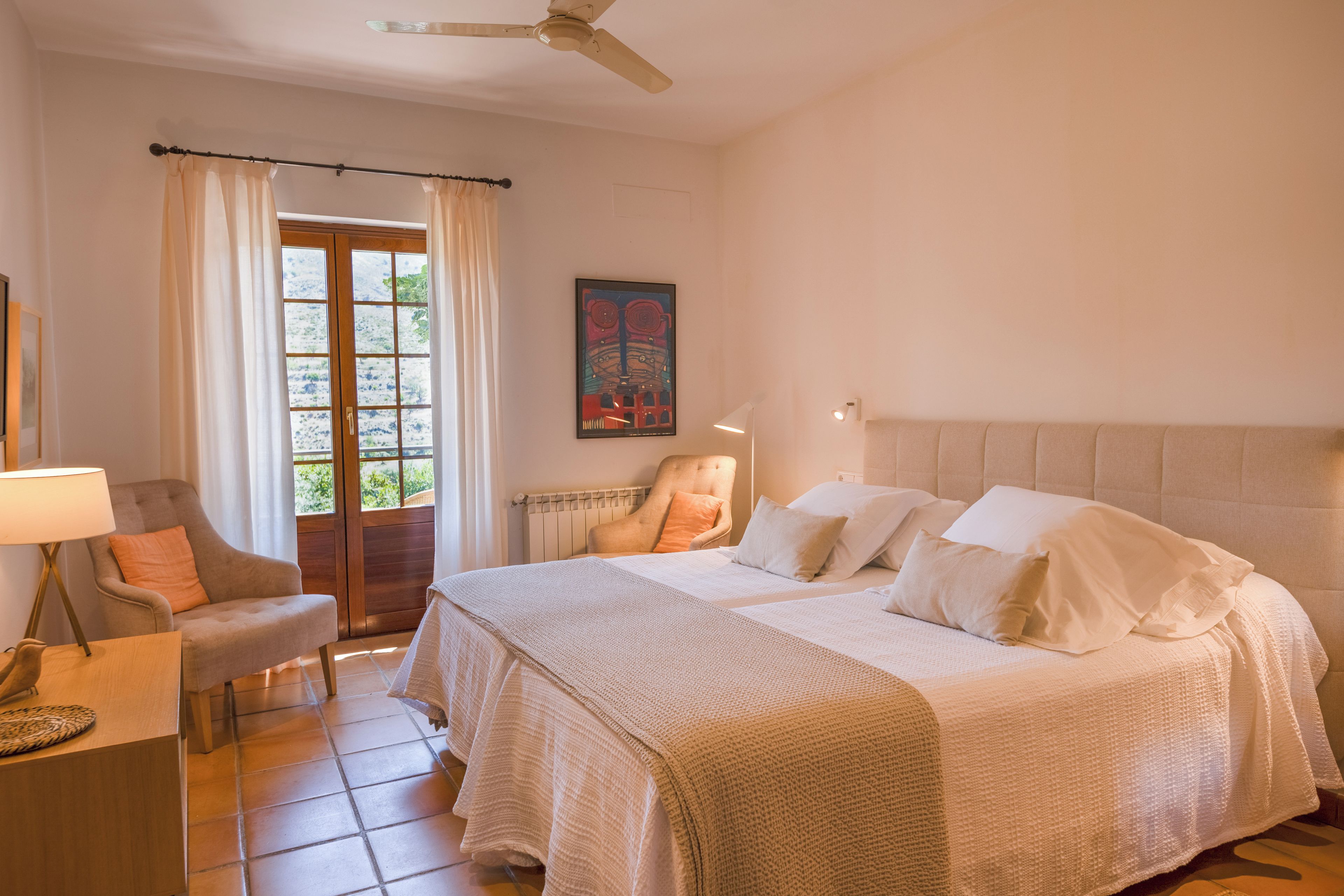 A bedroom with a double bed, a chair, a desk, and a window with curtains.