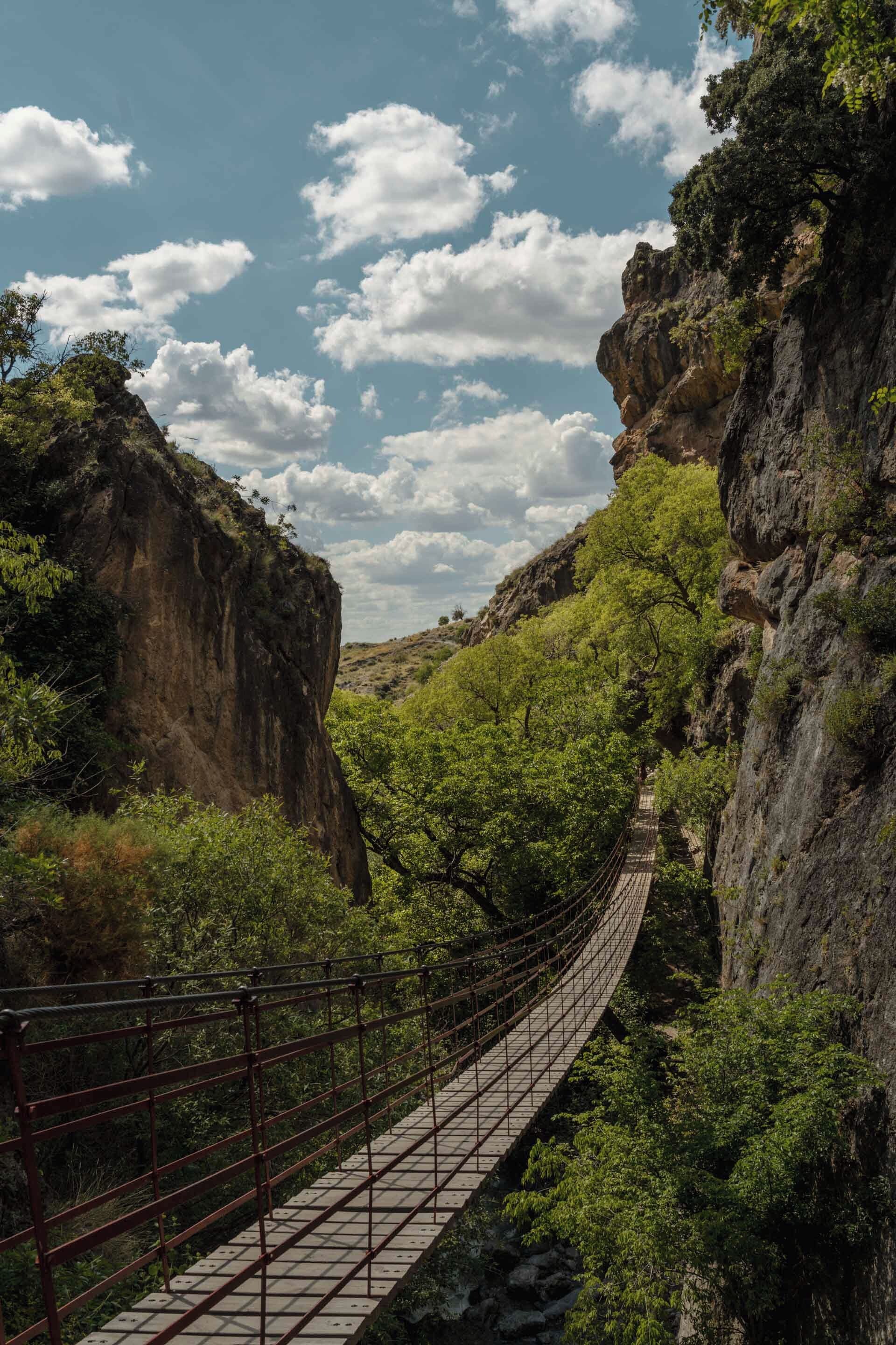 A suspension bridge in a mountainous area with rocky cliffs and dense greenery.