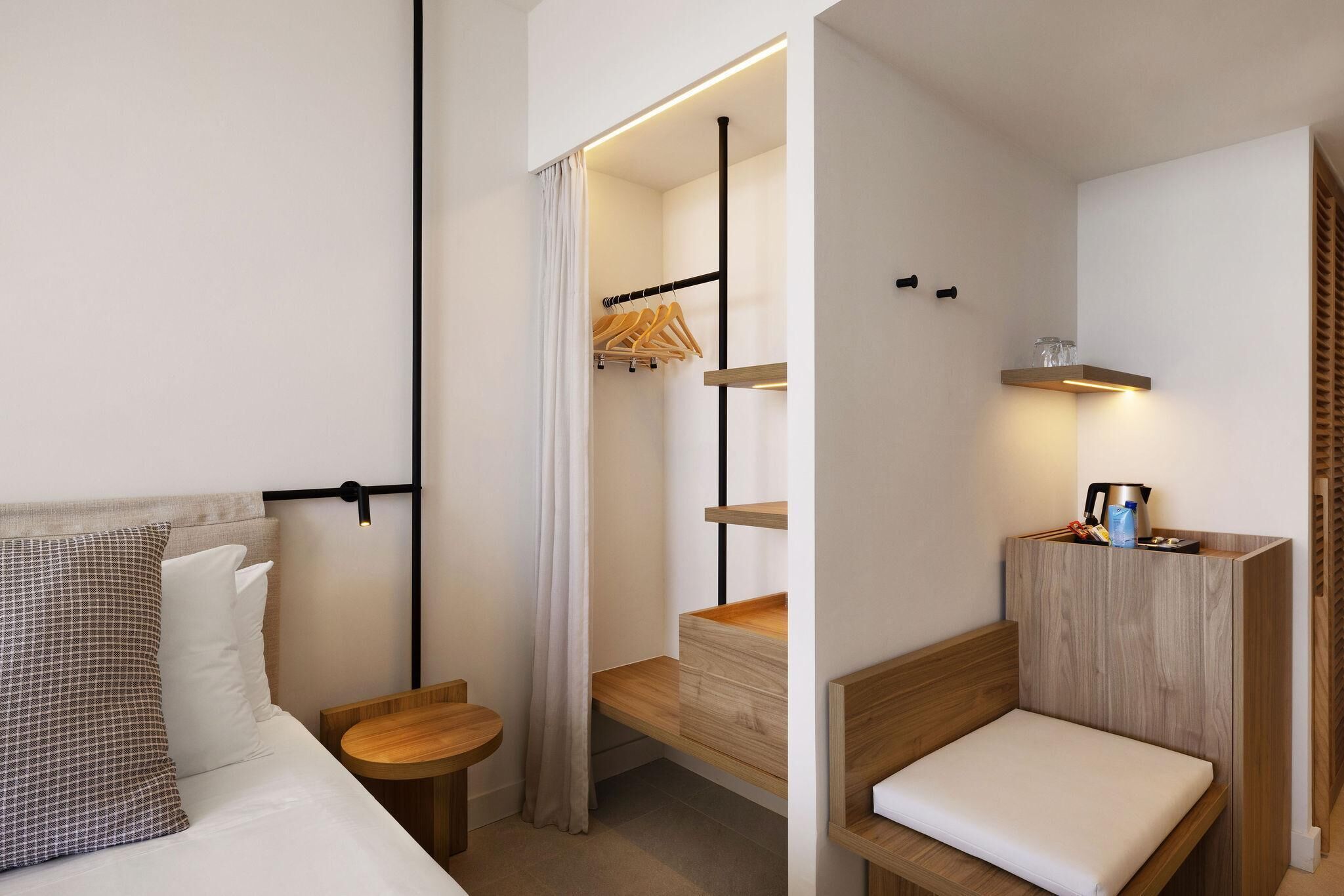 Habitación de hotel compacta con cama, mesita de madera, un banco empotrado con cojín, perchero y estante con hervidor y otros objetos.