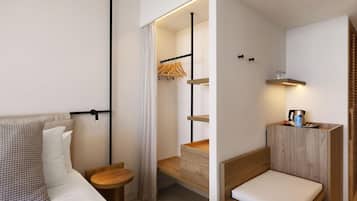 Habitación de hotel compacta con cama, mesita de madera, un banco empotrado con cojín, perchero y estante con hervidor y otros objetos.