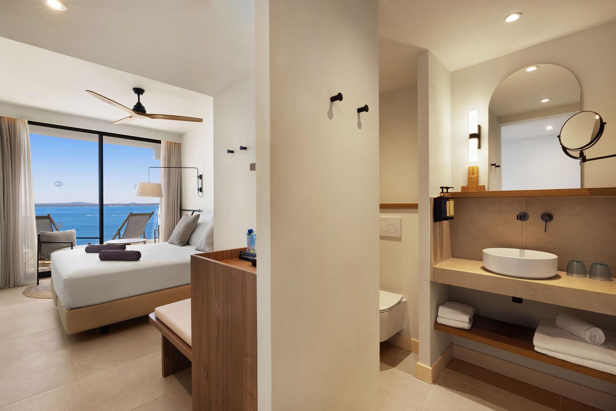 Une chambre d’hôtel moderne dotée d’un grand lit, offrant une vue sur la mer, comprenant une salle de bain avec un grand miroir rond et un ventilateur de plafond.
