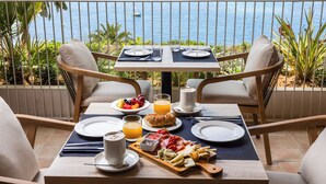 Una mesa lista para el desayuno con vista al mar, que incluye platos, vasos y una bandeja de frutas.