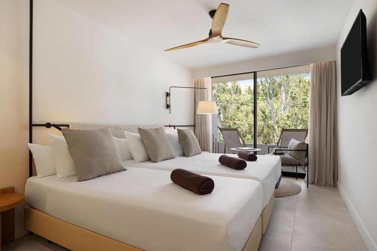 A hotel room with two beds, a ceiling fan, a TV, a window with a view of trees, and a desk with a chair.