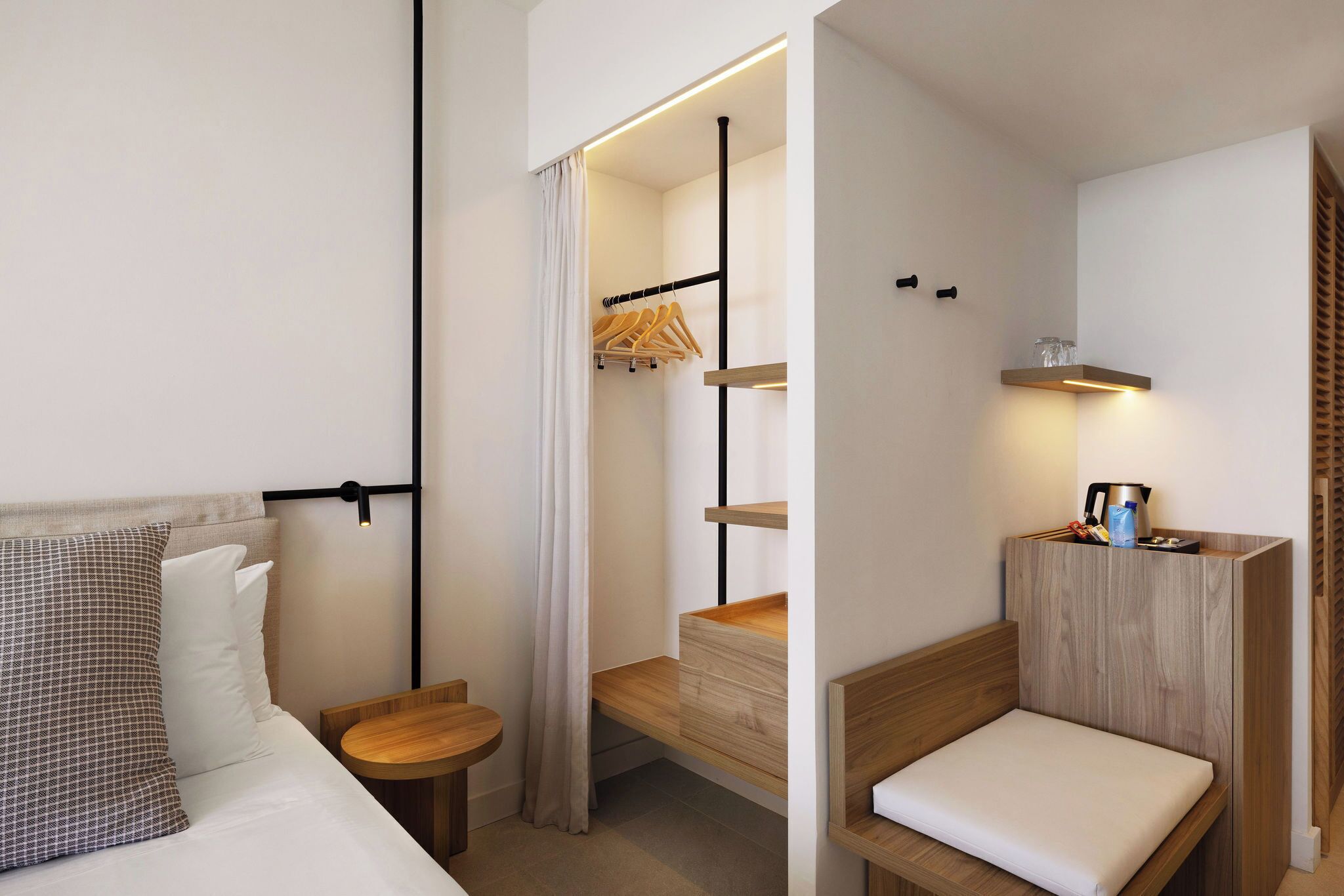 Et moderne hotelværelse med en seng, et lille trænatbord, en indbygget bænk, et knagered og en hylde med elkedel og andet udstyr.