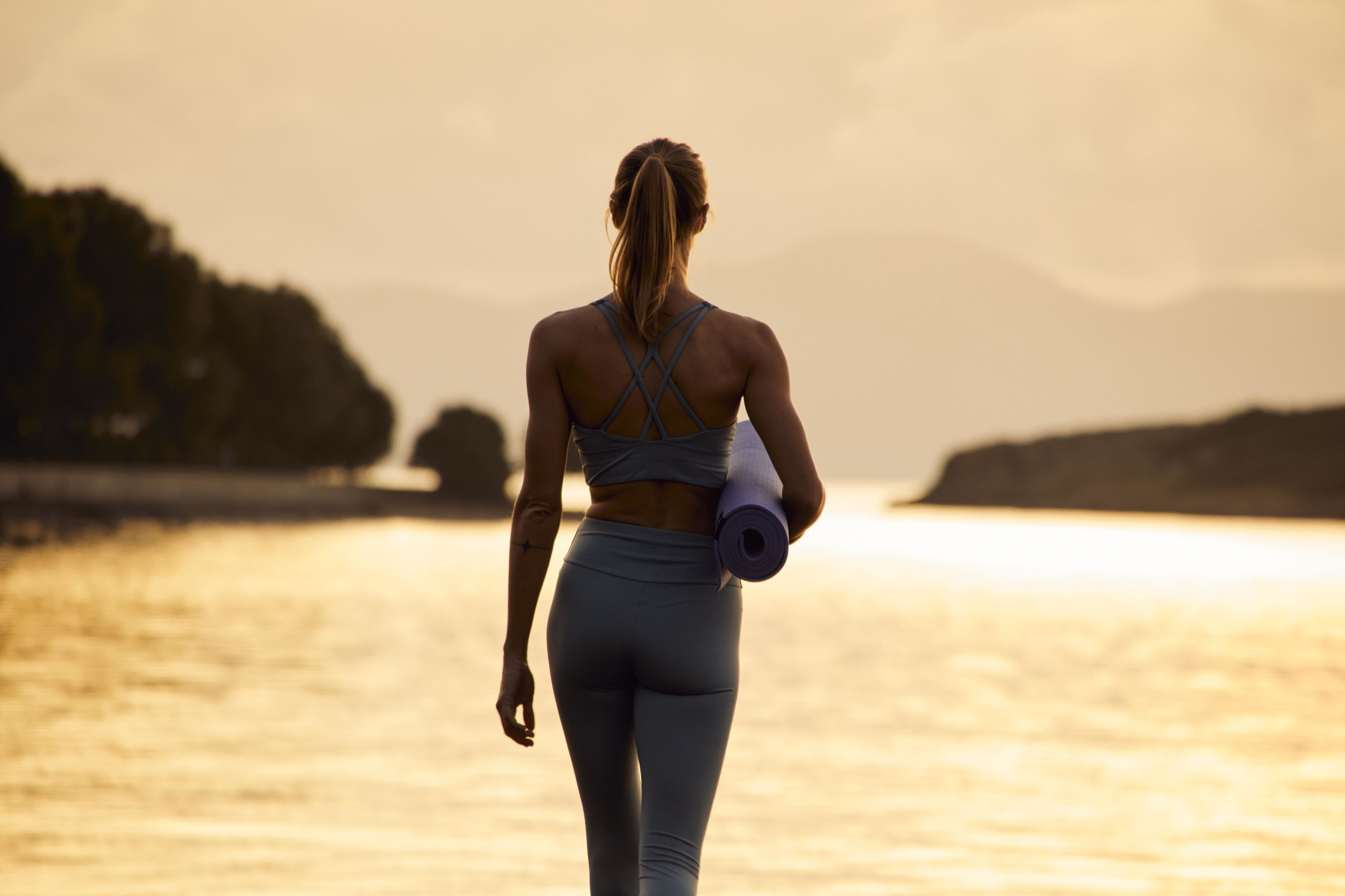 Una persona caminando sobre la playa con una esterilla de yoga, usando un top deportivo y leggings.