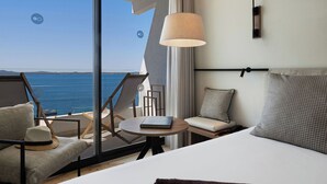 Habitación de hotel con un ventanal grande con vista al mar, una cama, una mesa redonda con un libro y una silla con un sombrero.