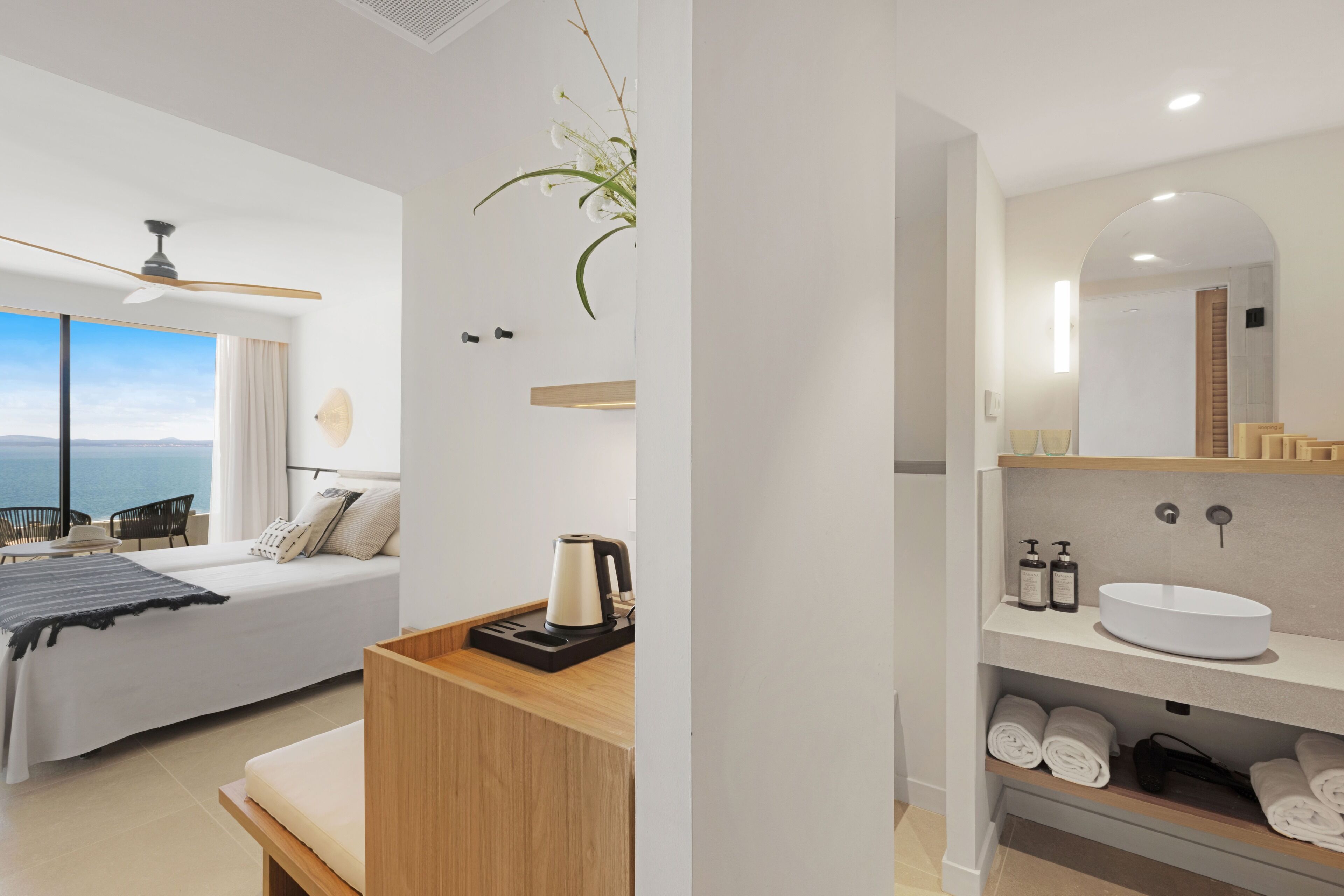 Una habitación de hotel moderna con una cama, una mesita de noche de madera, una cafetera, un ventanal con vistas y un baño con lavabo y toallas.