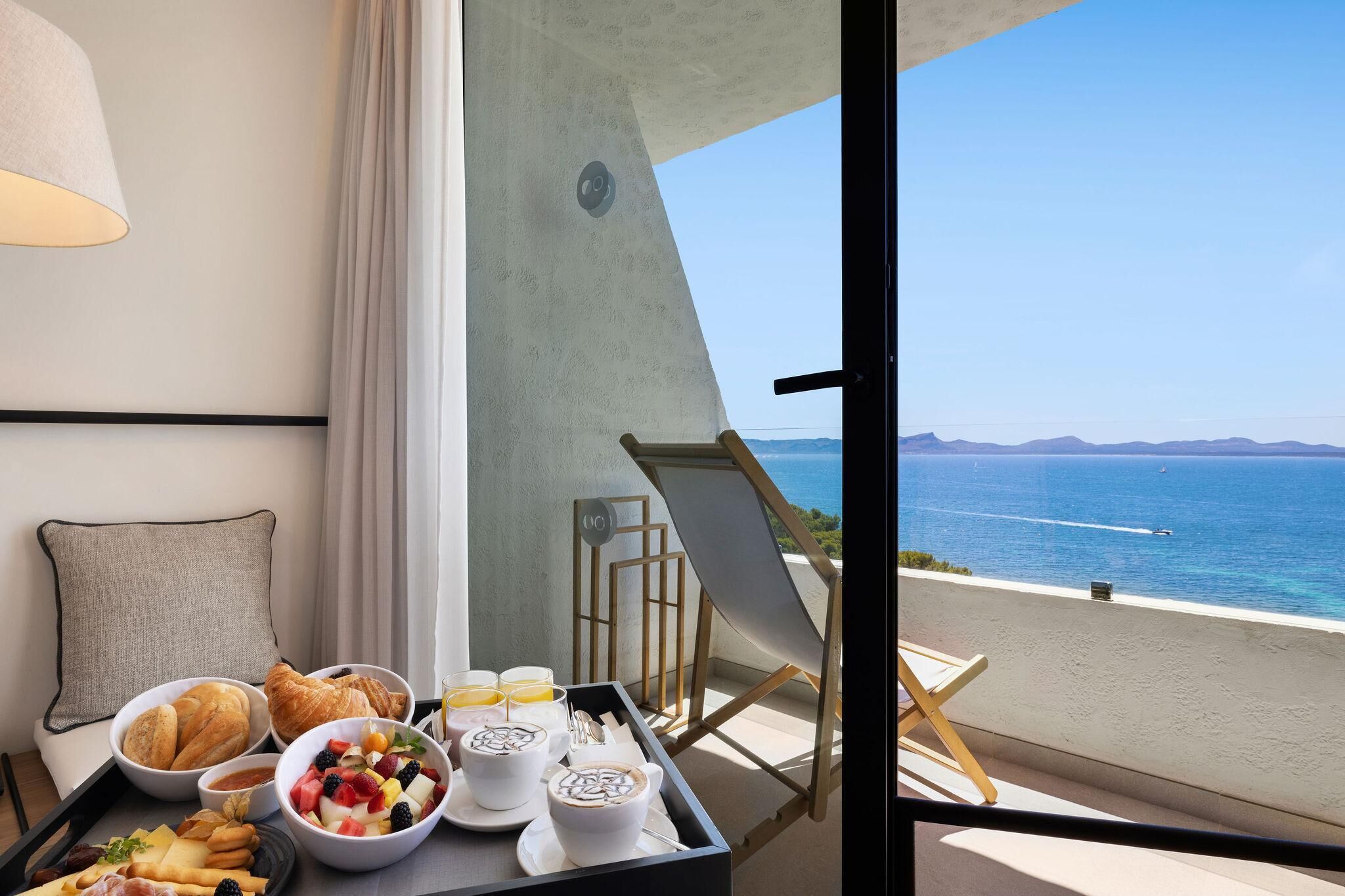 Un balcón con comedor que incluye cruasanes, fruta y café, con vistas a la costa, barcos y montañas.
