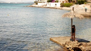 Escena costera con una mujer de pie sobre una plataforma de concreto frente al mar, observando una hilera de edificios blancos con balcones y árboles.
