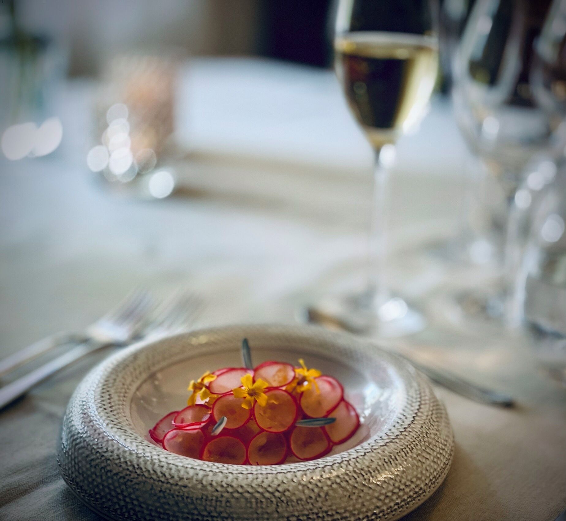 Ett matbord med en vävd tallrik som innehåller röda vindruvor och gula blommor, ett glas vitt vin och en suddig bakgrund med ljus.