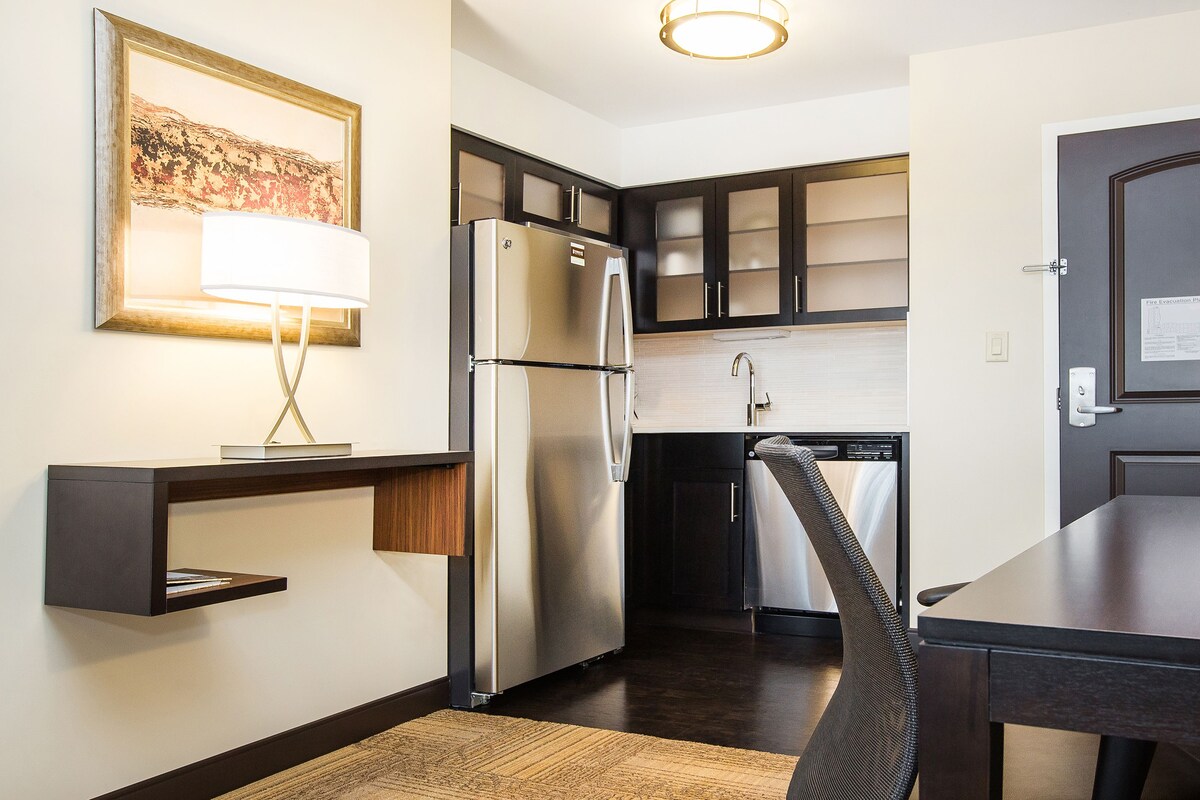 A compact kitchen with a refrigerator, stove, and microwave. There is a chair and a small table. A fire evacuation plan is posted on the door.