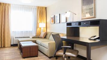 Habitación de hotel con escritorio, una silla, un sofá, una lámpara, teléfono y ventana con cortinas.