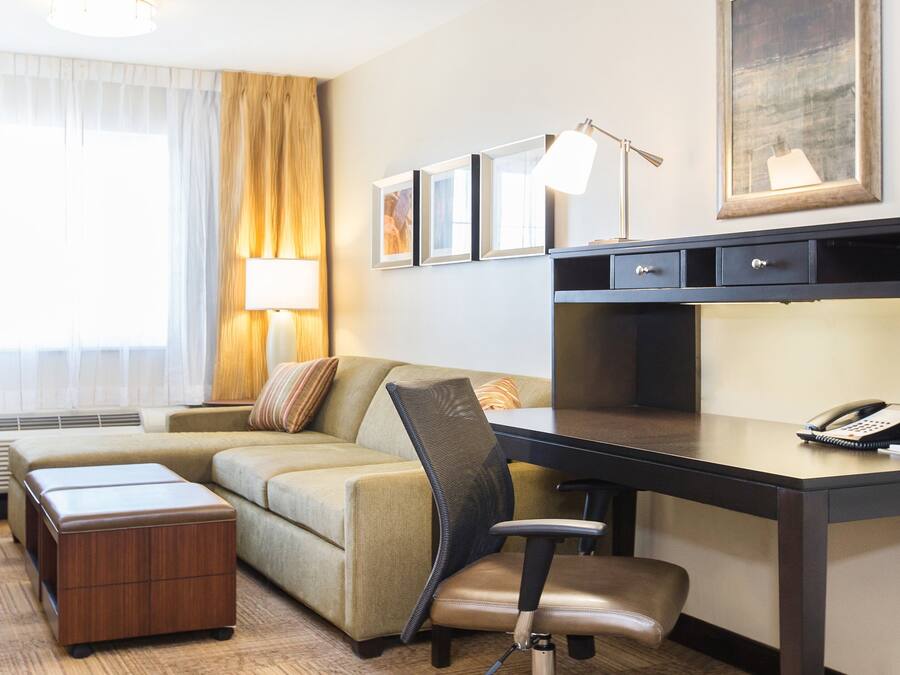 Habitación de hotel con escritorio, una silla, un sofá, una lámpara, teléfono y ventana con cortinas.