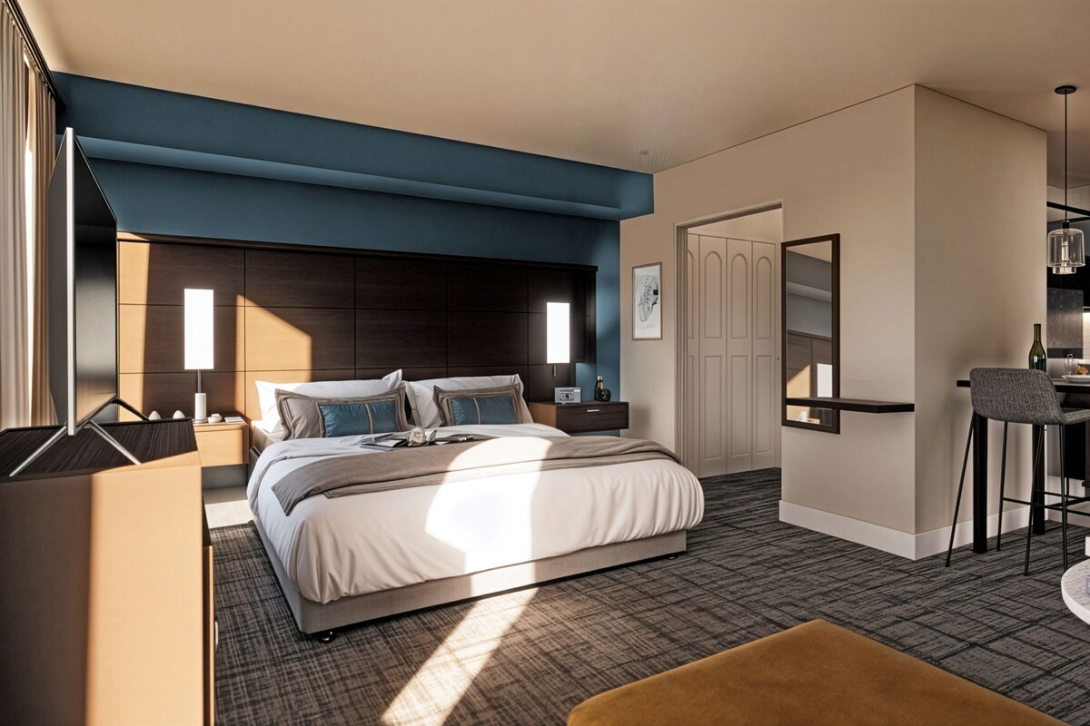 A modern hotel room with a large bed, a desk, a chair, a small table, and a potted plant.
