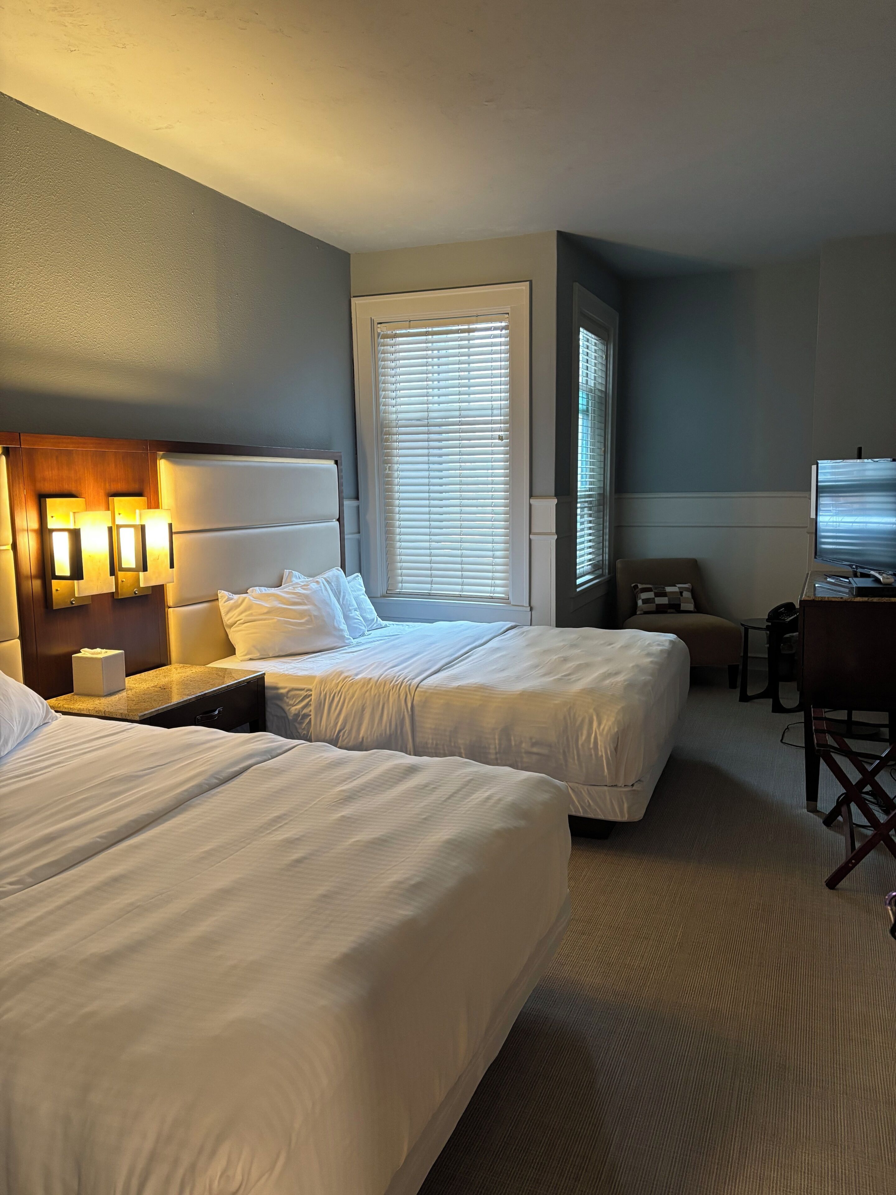 A hotel room with two beds, a desk, a chair, and a television.