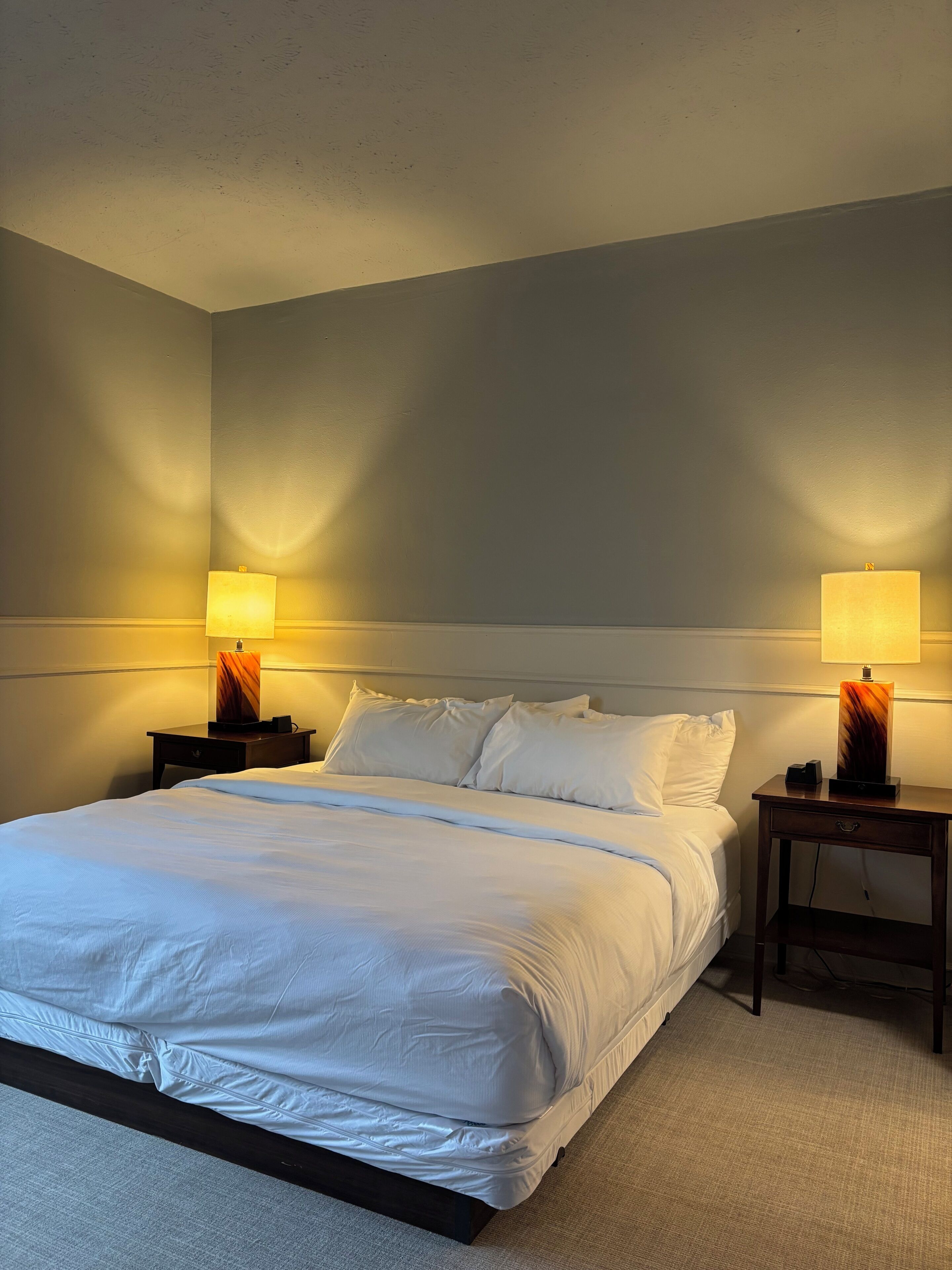 A hotel room with a large bed, two bedside tables with lamps, and a plain wall.