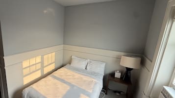 A hotel room with a bed, bedside table, lamp, and a window with blinds.