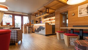 Un lobby de hotel acogedor con pisos de madera, una zona de bar con estantes llenos de botellas, y un ventanal con vistas al exterior.
