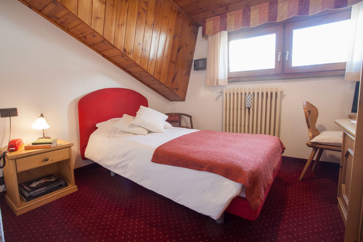 Une chambre à coucher avec un lit rouge, une table de chevet en bois avec une lampe, une chaise et une fenêtre avec des rideaux.