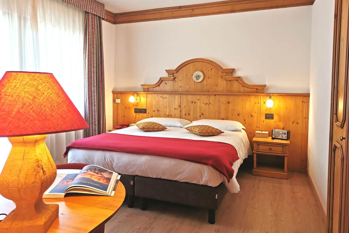 Une chambre d’hôtel avec une tête de lit en bois, un lit recouvert d’une couverture rouge, une table de chevet avec une lampe et un livre, et une petite table de nuit avec une radio.