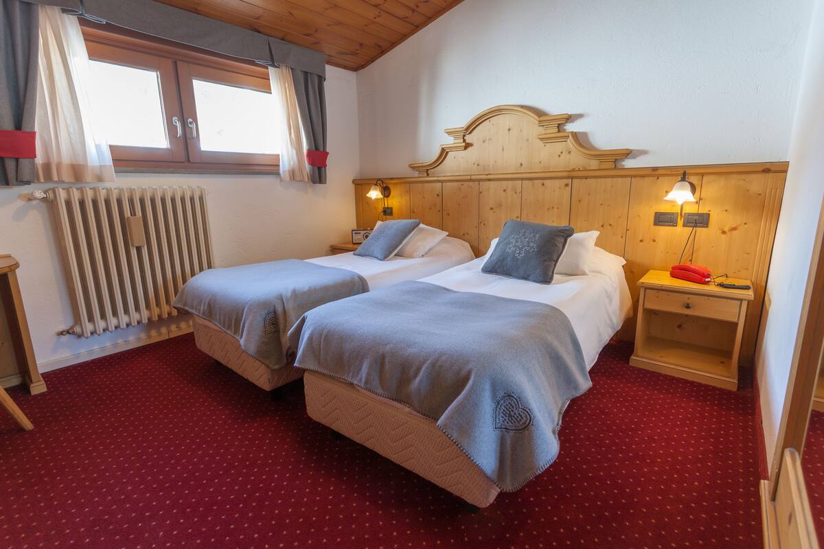 Une chambre double avec deux lits, une tête de lit en bois, une moquette rouge, un radiateur et une fenêtre avec des rideaux.