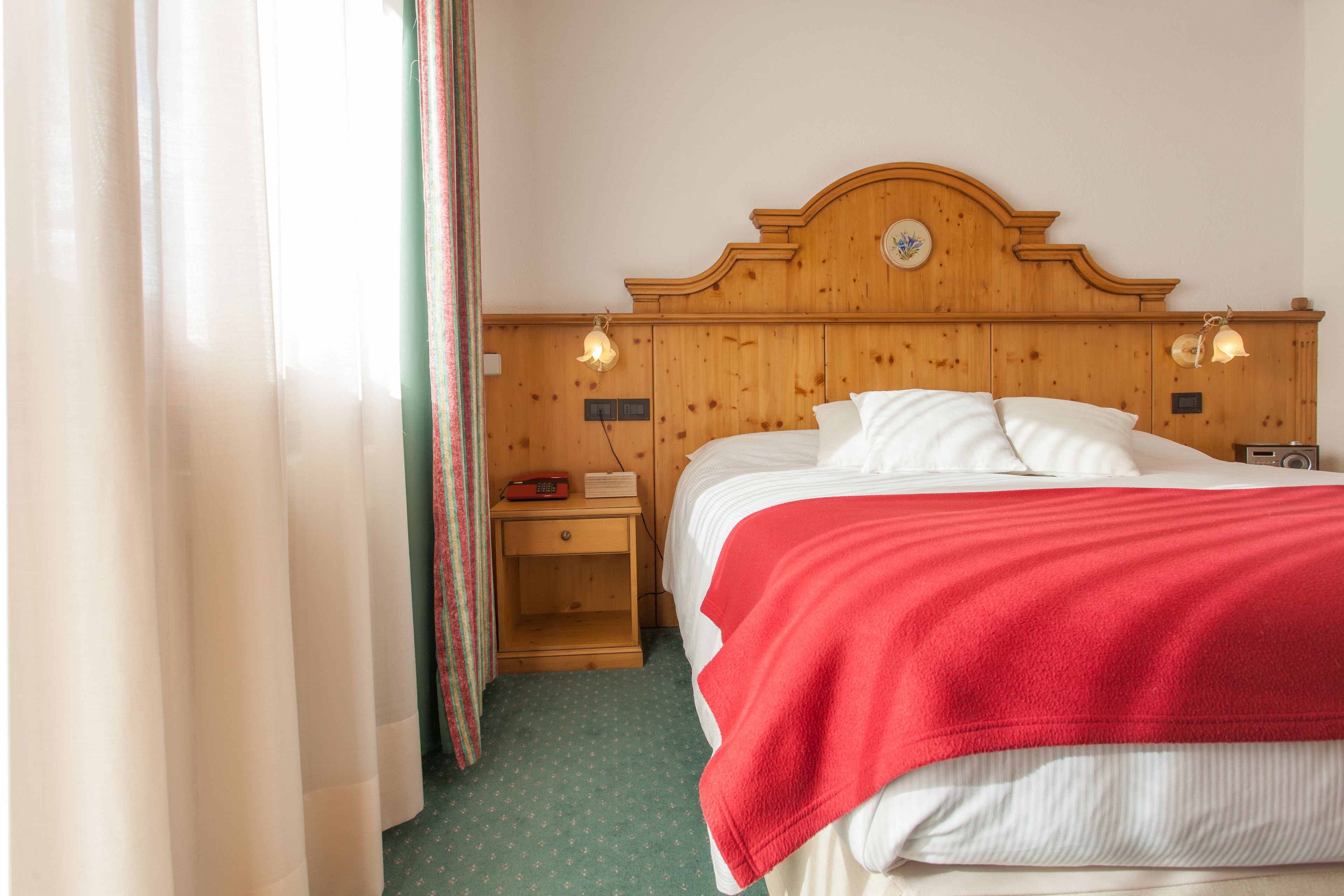 Une chambre d’hôtel avec une tête de lit en bois, un lit avec une literie blanche et une couverture rouge, une table de chevet avec une lampe, et une fenêtre avec des rideaux.