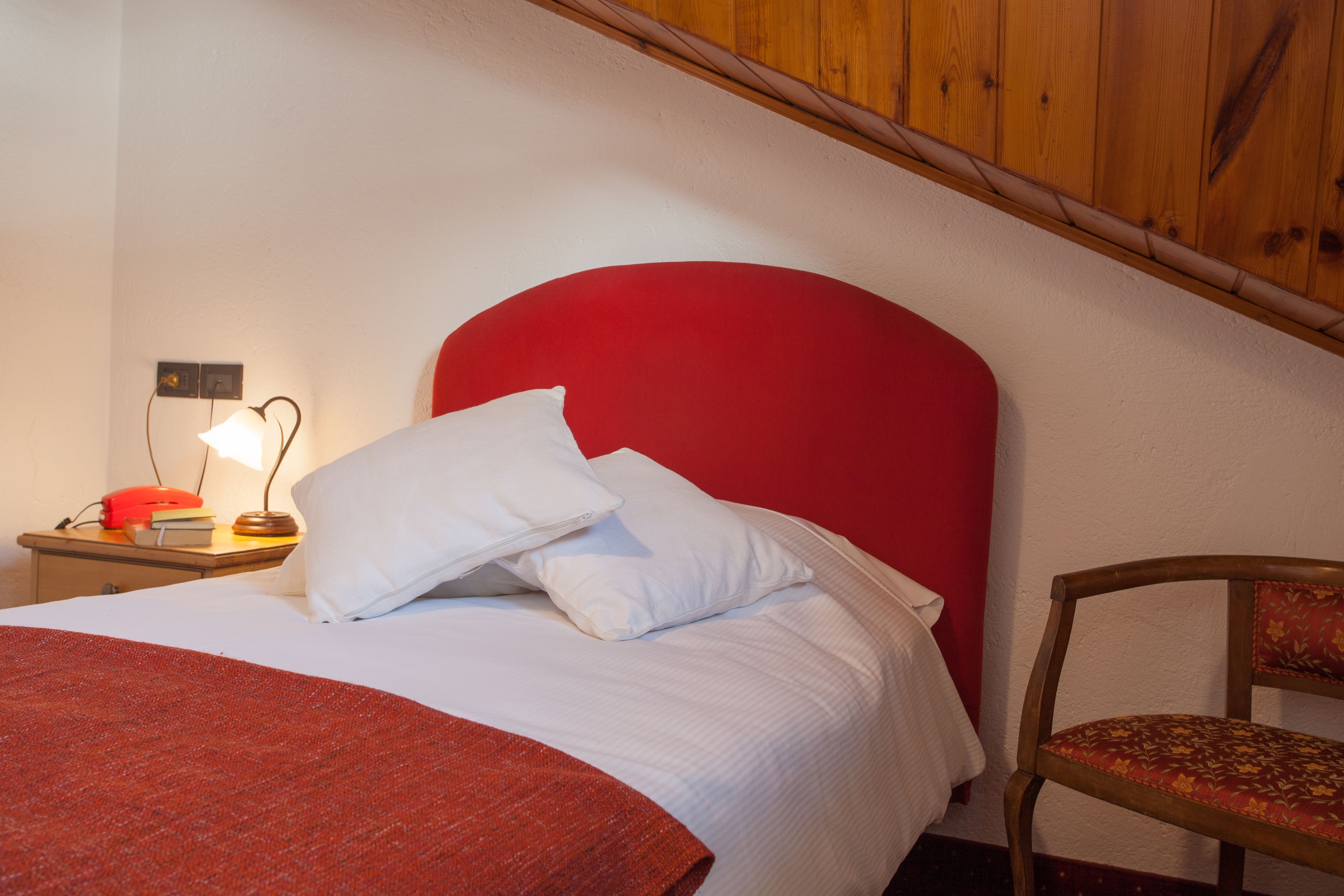 A bed with a red headboard, white pillows, and a red blanket. There is a wooden nightstand with a lamp and a telephone. A wooden chair with a floral cushion is visible.