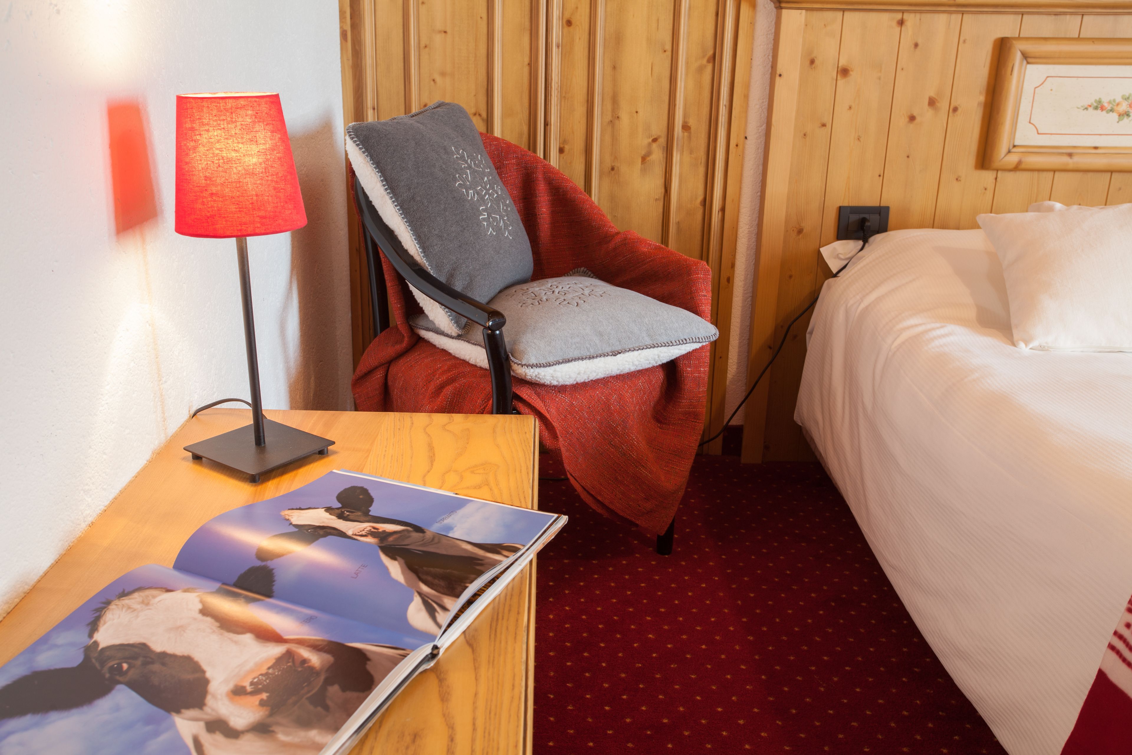 A hotel room with a wooden headboard, a red chair with a grey cushion, a white bed, and a wooden nightstand with a lamp and a magazine.