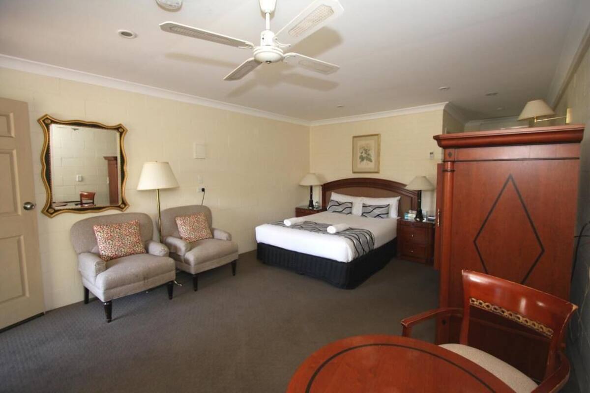 A hotel room with a bed, two armchairs, a wooden cabinet, a ceiling fan, and a mirror on the wall.