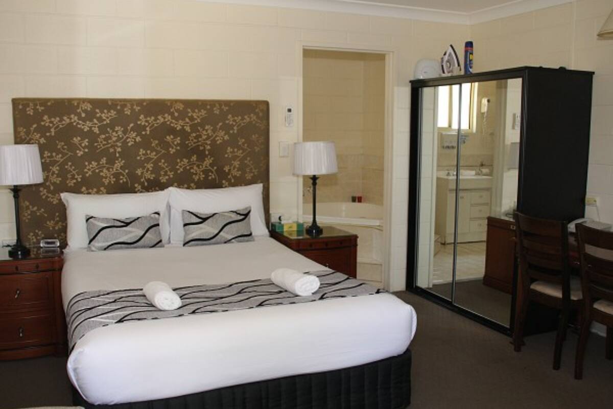 A hotel room with a large bed, two bedside tables, a desk, a chair, a bathroom with a glass shower enclosure, and a wardrobe.