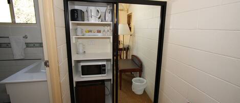 A compact kitchen with a microwave, shelves with kitchenware, a small table, and a trash bin.