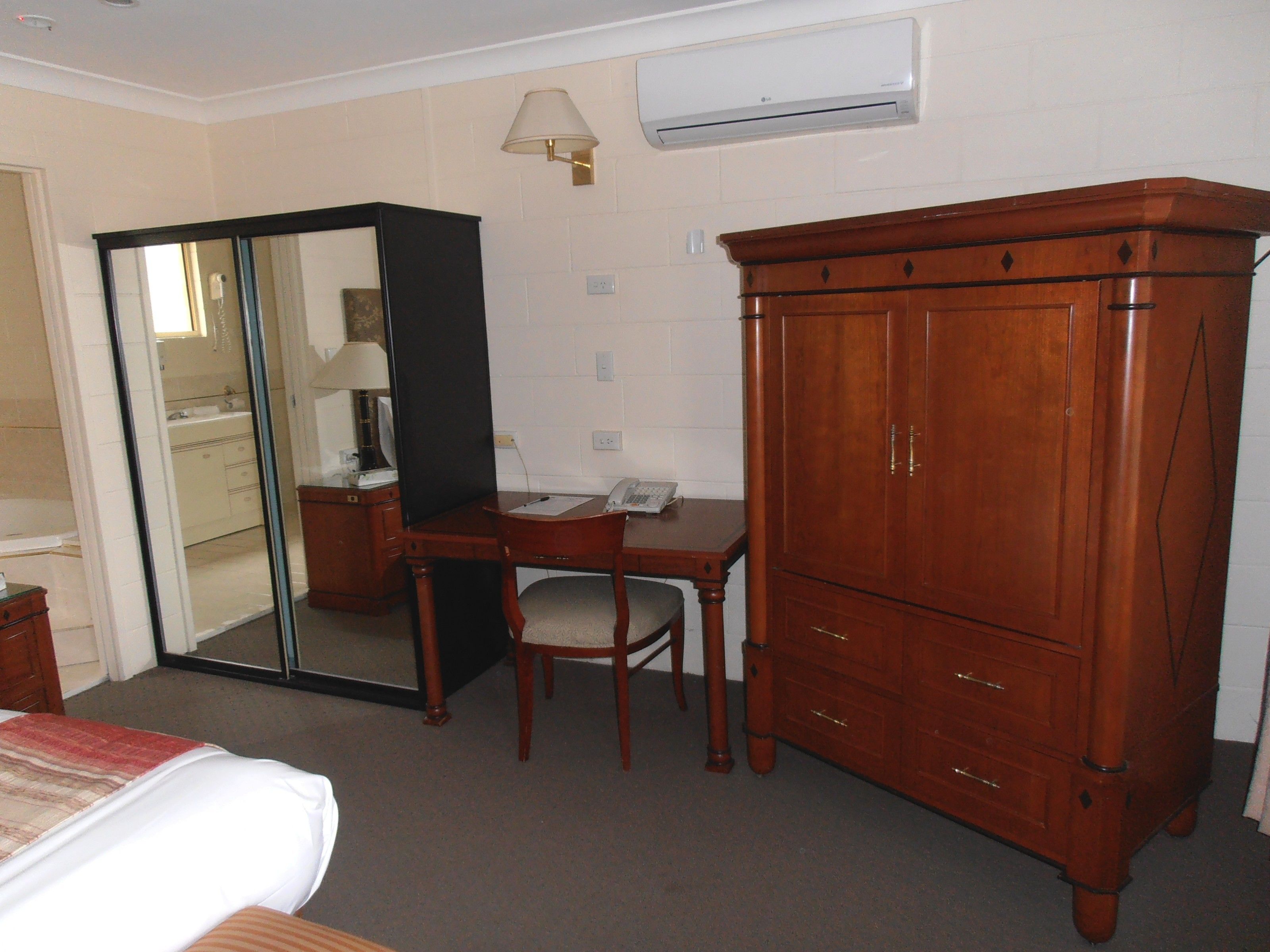 A hotel room with a bed, a desk, a chair, a wardrobe, a mirror, and an air conditioning unit.