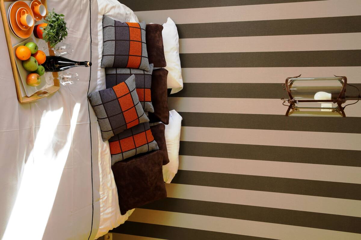 A neatly arranged bed with striped bedding, a bedside table with a lamp, and a tray with fruits and a bottle.