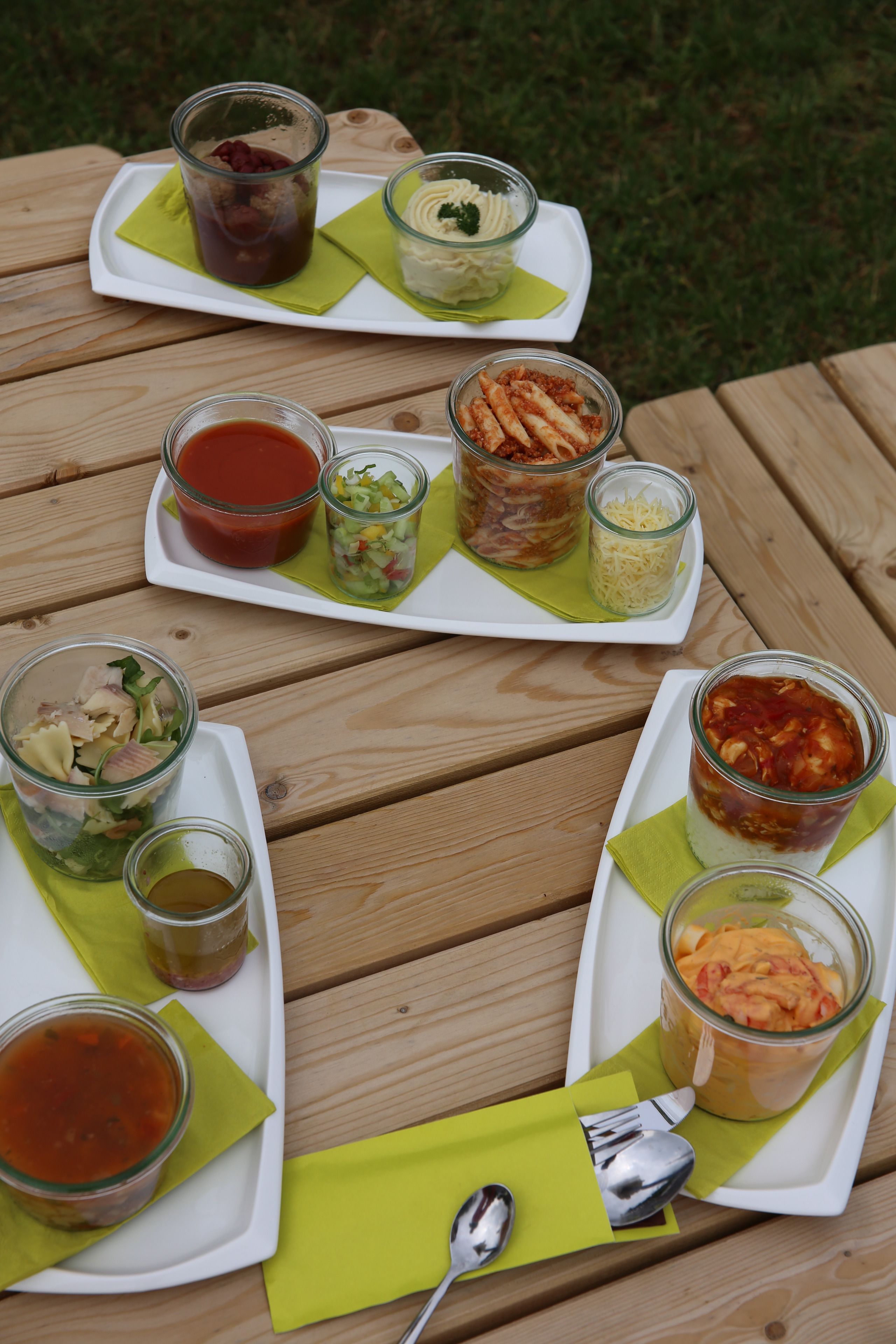 A wooden table with various food items and condiments arranged on white plates and glass jars, accompanied by green napkins and a spoon.