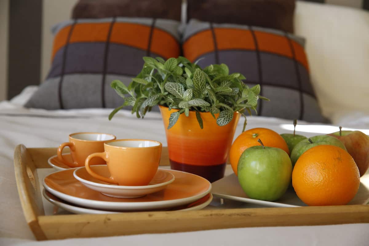 A breakfast tray with orange cups, plates, a glass of orange juice, apples, and a potted mint plant on a bed with orange and gray pillows.