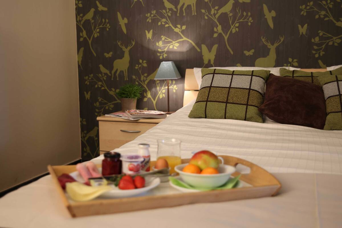A neatly made bed with a tray of breakfast items, a bedside lamp, and a nightstand with a plant and a magazine.