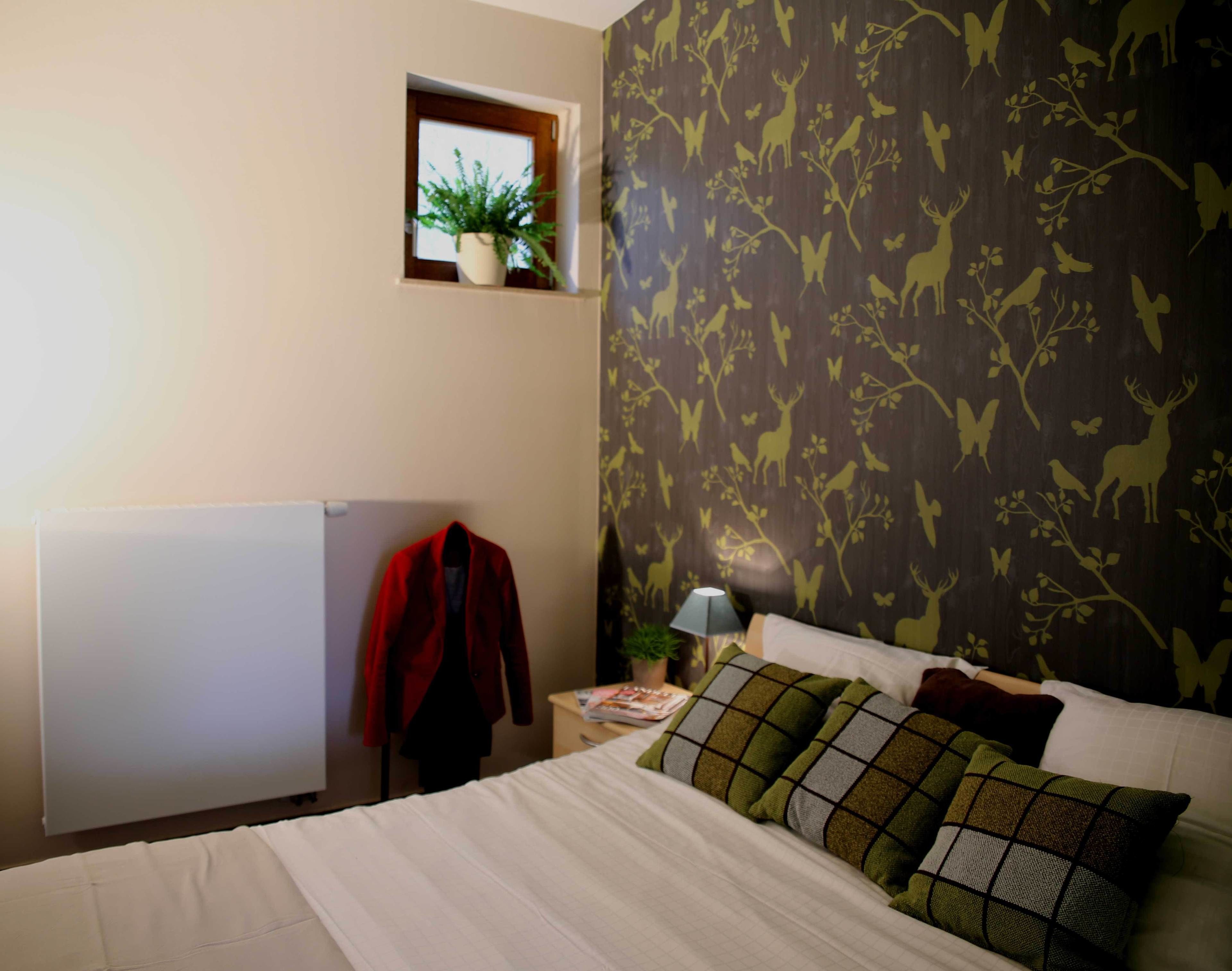 Une chambre à coucher comprenant un lit, une table de chevet, une lampe, une fenêtre avec une plante et un mur recouvert d’un papier peint à motifs naturels.